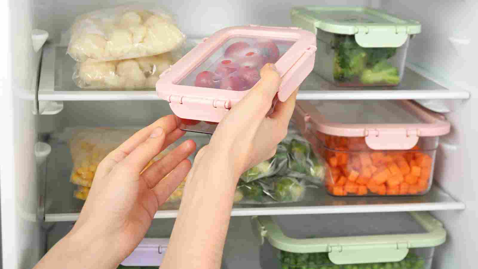 Hands placing a container of grapes in a refrigerator, with other containers of assorted vegetables and food items on the shelves.