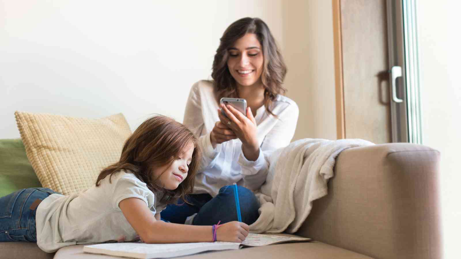 Mom scrolling on the phone while her child is drawing something on a book.