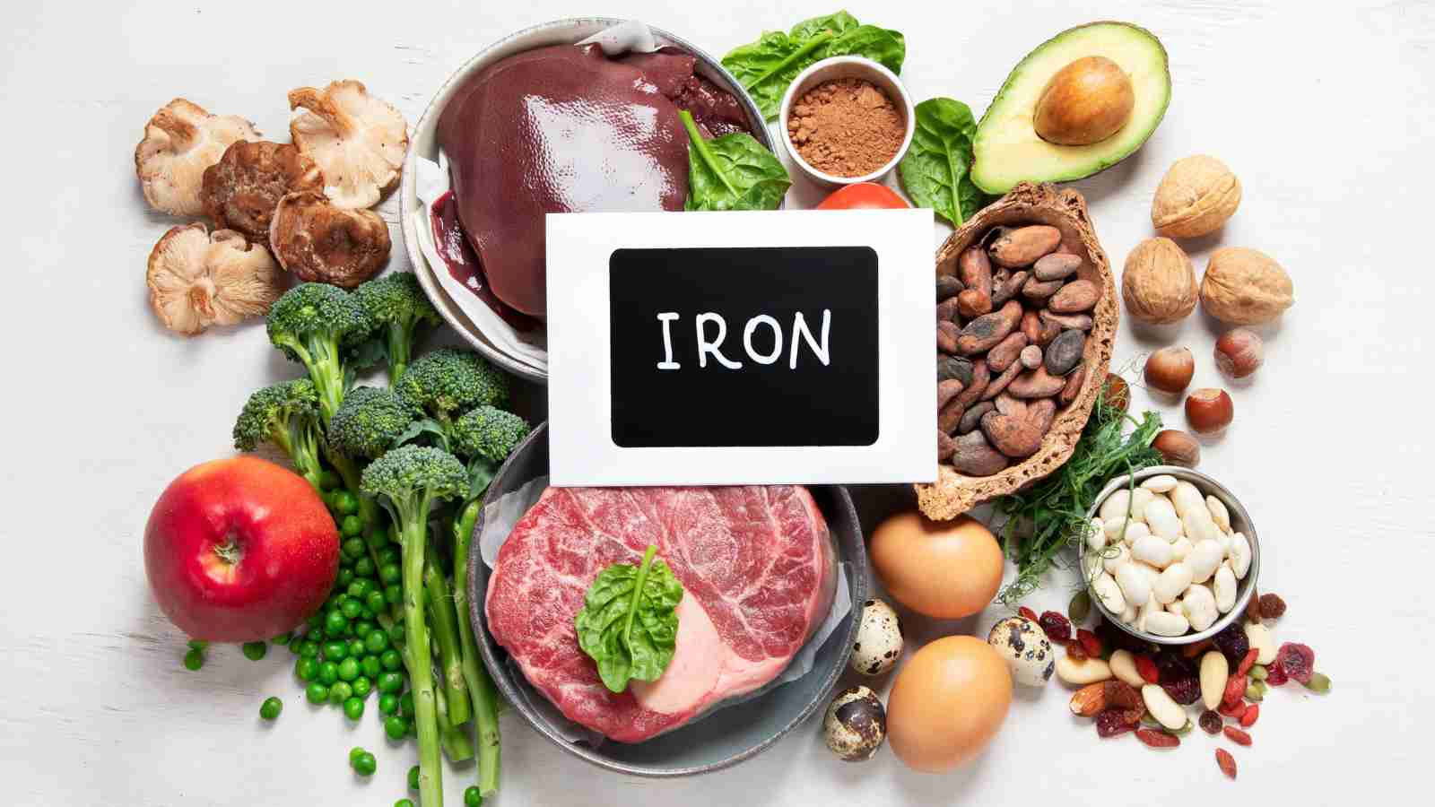 A variety of iron-rich foods, including meat, liver, vegetables, nuts, and seeds, are arranged around a small chalkboard with the word "IRON" written on it.