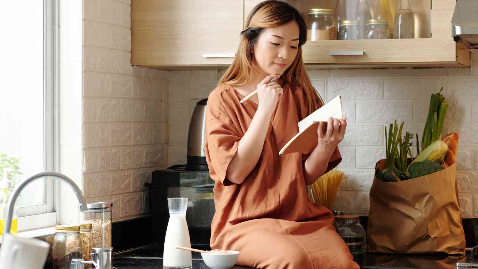 A woman sits on a kitchen counter writing in a notebook, with groceries, milk, and a bowl of food nearby.