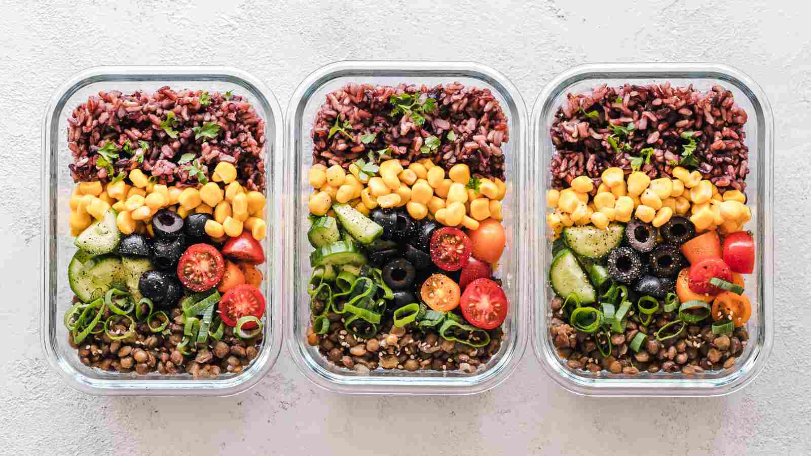 Three glass containers filled with layered salads, including brown rice, corn, cherry tomatoes, black olives, cucumber, green onions, and lentils, arranged in rows on a light surface.
