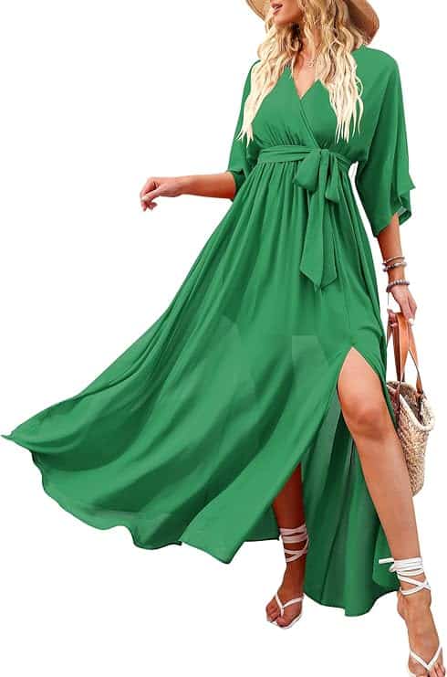 Woman wearing a green, flowy, V-neck maxi dress with a waist tie, high slit, and paired with strappy sandals, bracelets, and a woven bag. Her head is not shown.