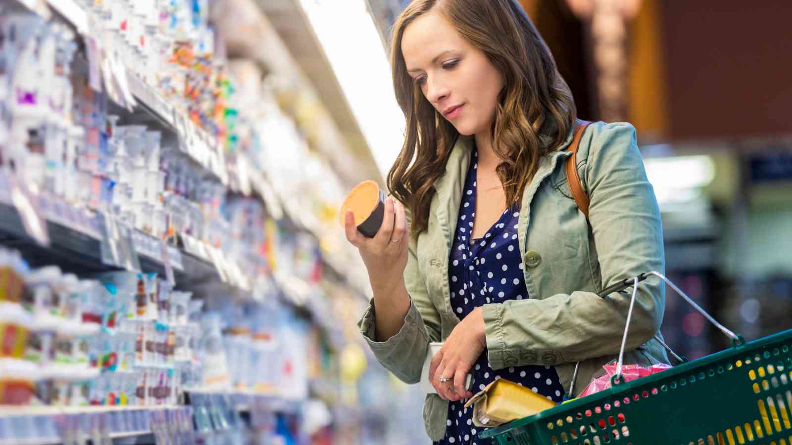 Woman holding a shopping basket examines a packaged food item in a grocery store aisle.