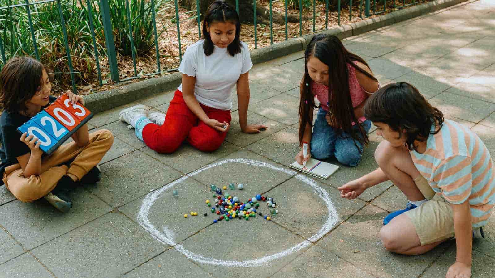 Four children sit on pavement around a chalk circle filled with marbles; one holds a notebook, another holds a scoreboard, and two appear to be playing marbles.