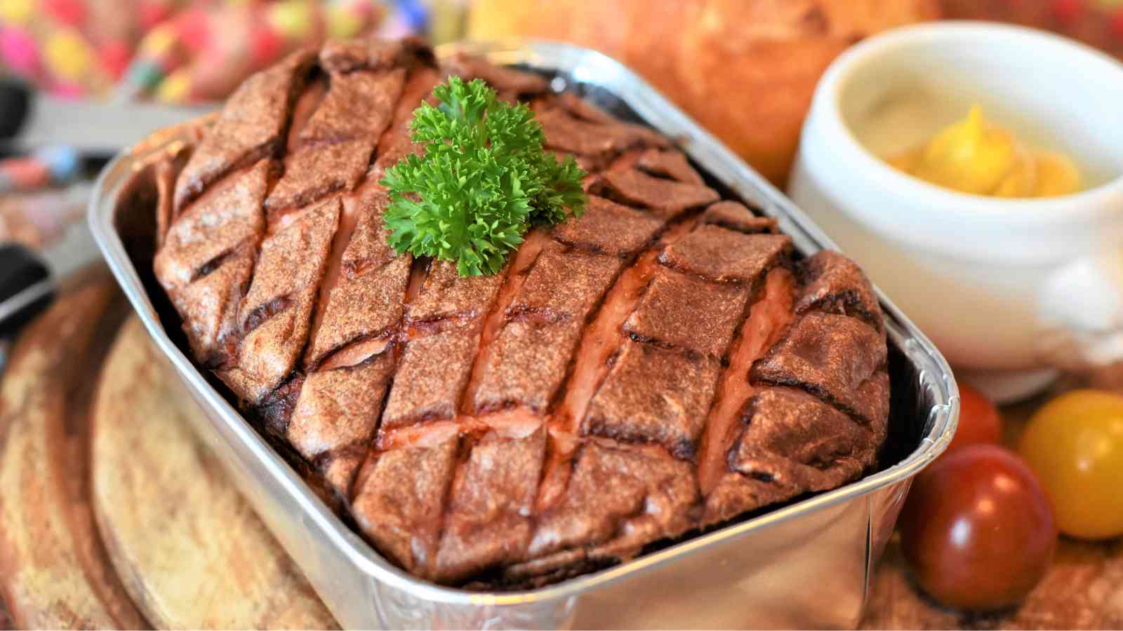 A baked meatloaf in a metal tray is garnished with parsley on top, with mustard, tomatoes, and bread visible in the background.
