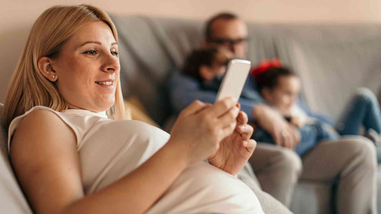 A pregnant woman sits on a couch using her smartphone, while a man and two children are seated in the background.