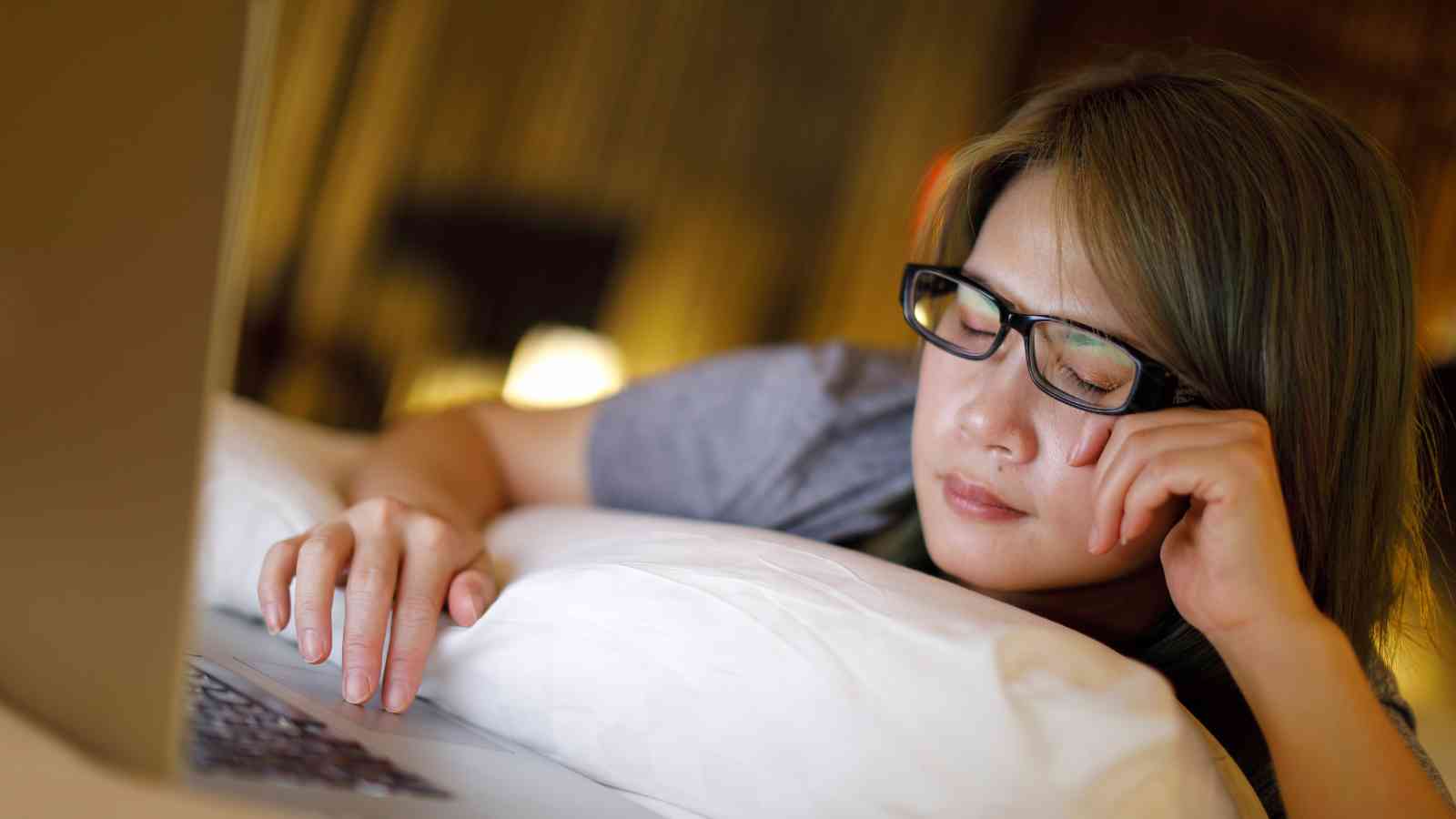 Person wearing glasses resting their head on a pillow, appearing to fall asleep while using a laptop.