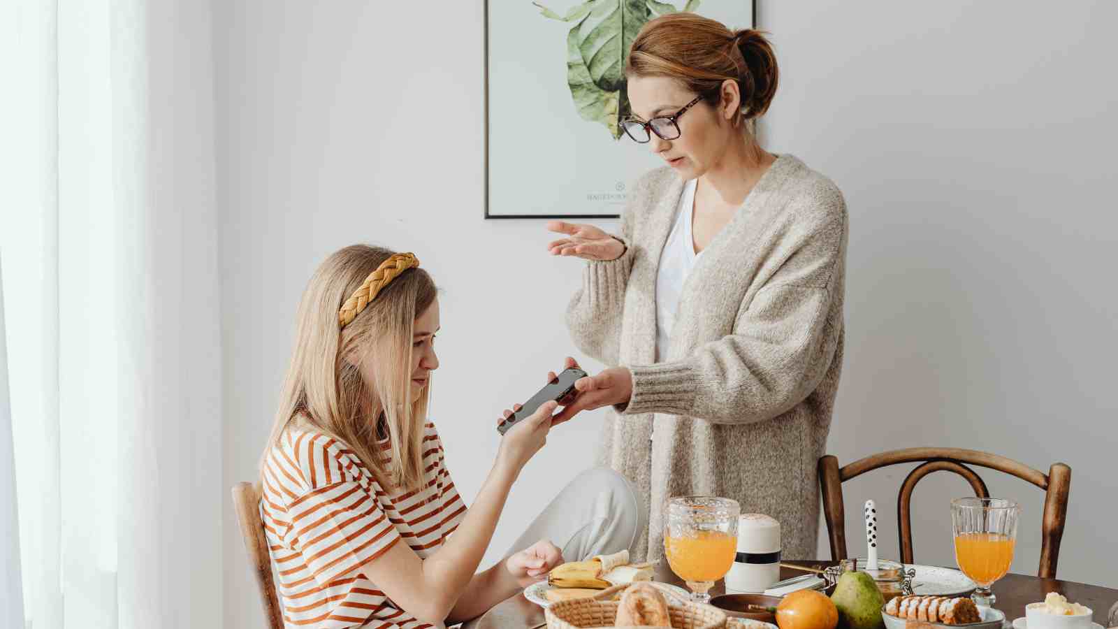 Two women sit at a dining table with breakfast items; one hands a phone to the other, who appears to be questioning or gesturing toward her.