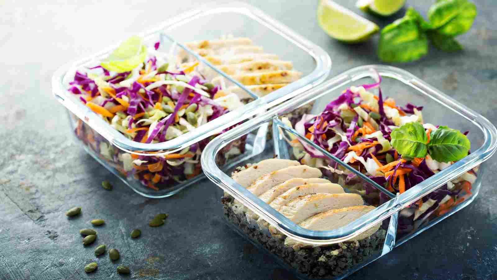 Two glass meal prep containers with sections holding sliced chicken breast, mixed vegetable slaw, and grains on a grey surface. Lime wedges and basil leaves in the background.
