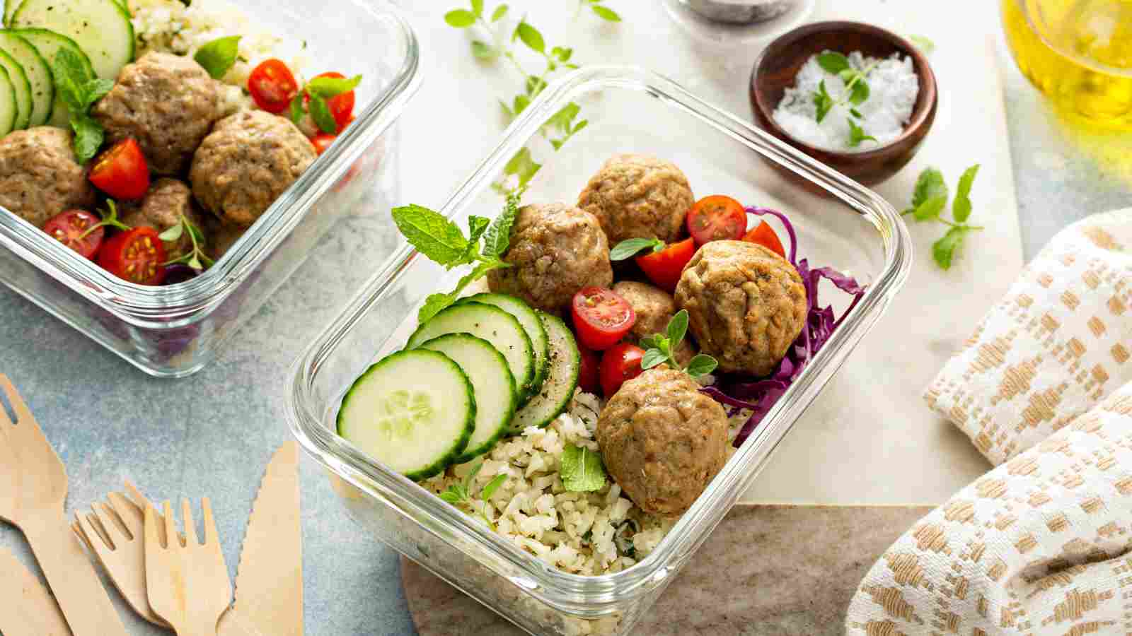 Glass meal prep containers with rice, meatballs, sliced cucumber, cherry tomatoes, and shredded cabbage, garnished with fresh herbs. Wooden utensils and salt in small bowl nearby.