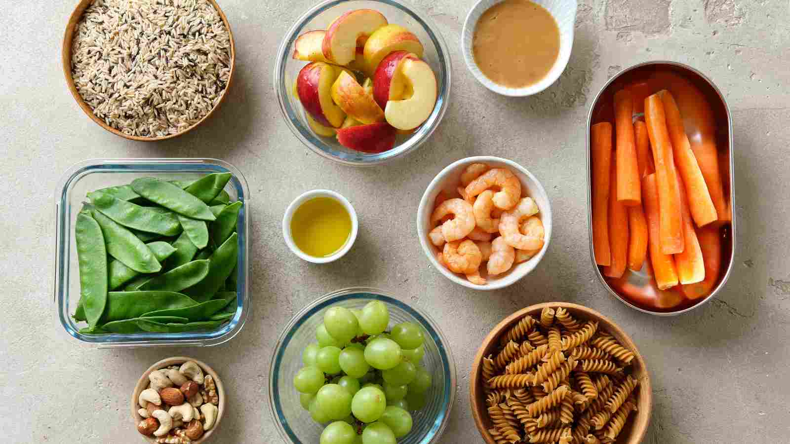 A variety of foods including cooked rice, sliced apples, peanut butter, carrot sticks, shrimp, snap peas, olive oil, green grapes, pasta, and mixed nuts arranged in bowls and containers.