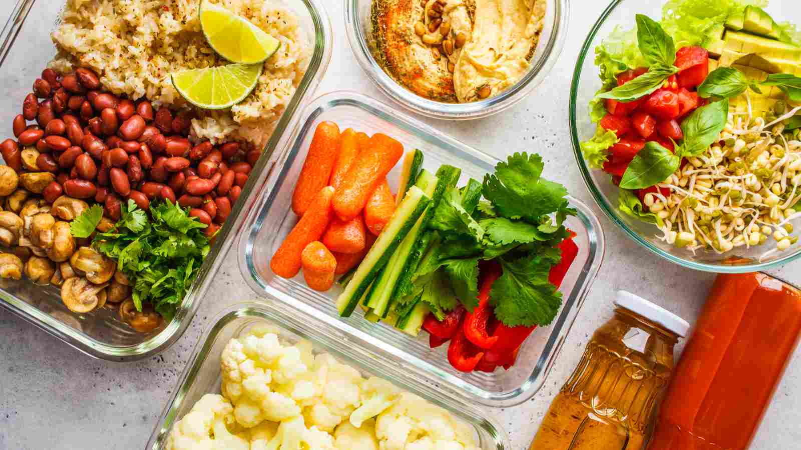 Assorted meal prep containers with beans, rice, chopped vegetables, salad, hummus, herbs, and two bottles of dressing arranged on a light surface.