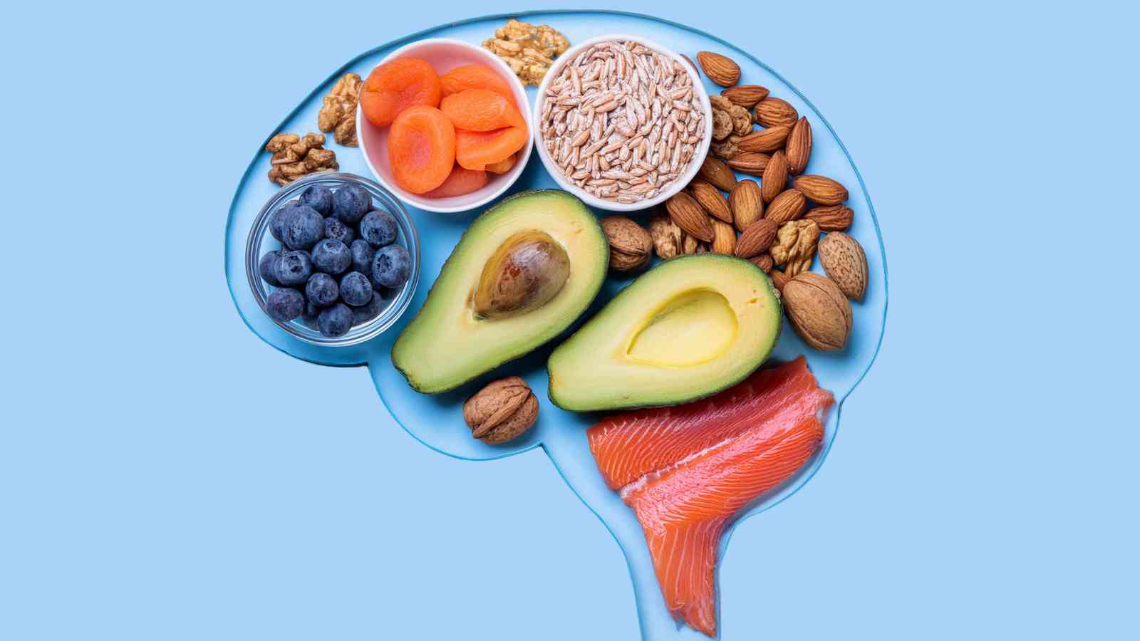 Brain-shaped arrangement of healthy foods, including avocados, blueberries, nuts, salmon, and dried apricots, on a light blue background.