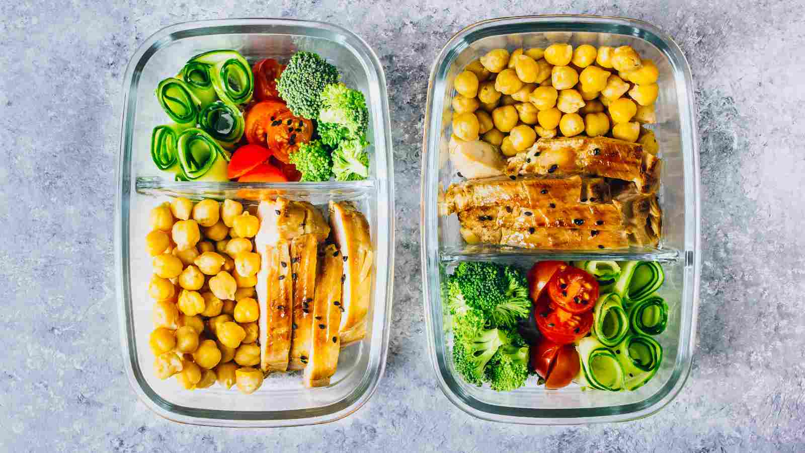 Two glass meal prep containers filled with sliced chicken, chickpeas, broccoli, sliced cucumber, and cherry tomatoes, arranged in divided sections.