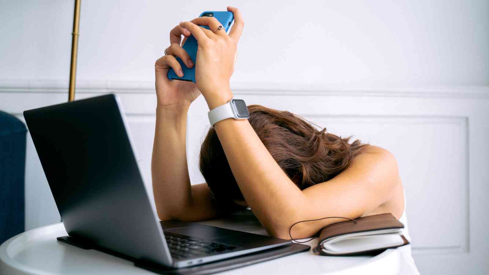 A person sits at a table with their head on their arm, holding a phone above their head next to an open laptop and a closed notebook.