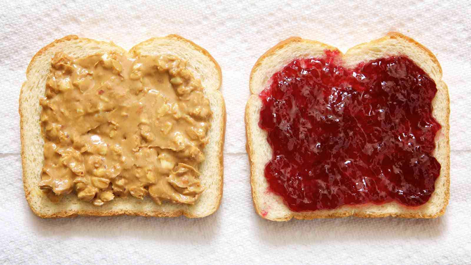Two slices of bread on a napkin, one spread with chunky peanut butter and the other with red grape jelly.