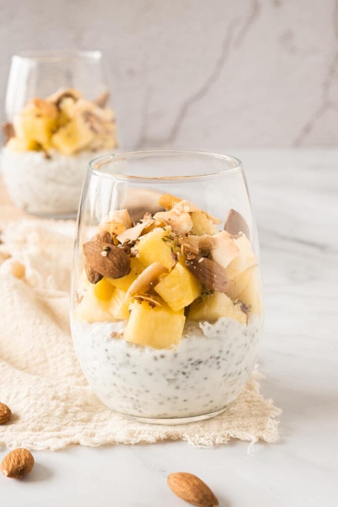 A glass filled with chia seed pudding topped with chopped pineapple, nuts, and seeds, placed on a light cloth with a second glass in the background.