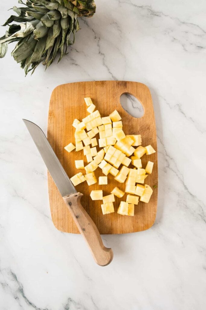 A wooden cutting board with diced pineapple pieces and a knife, with a pineapple crown visible in the top left corner.