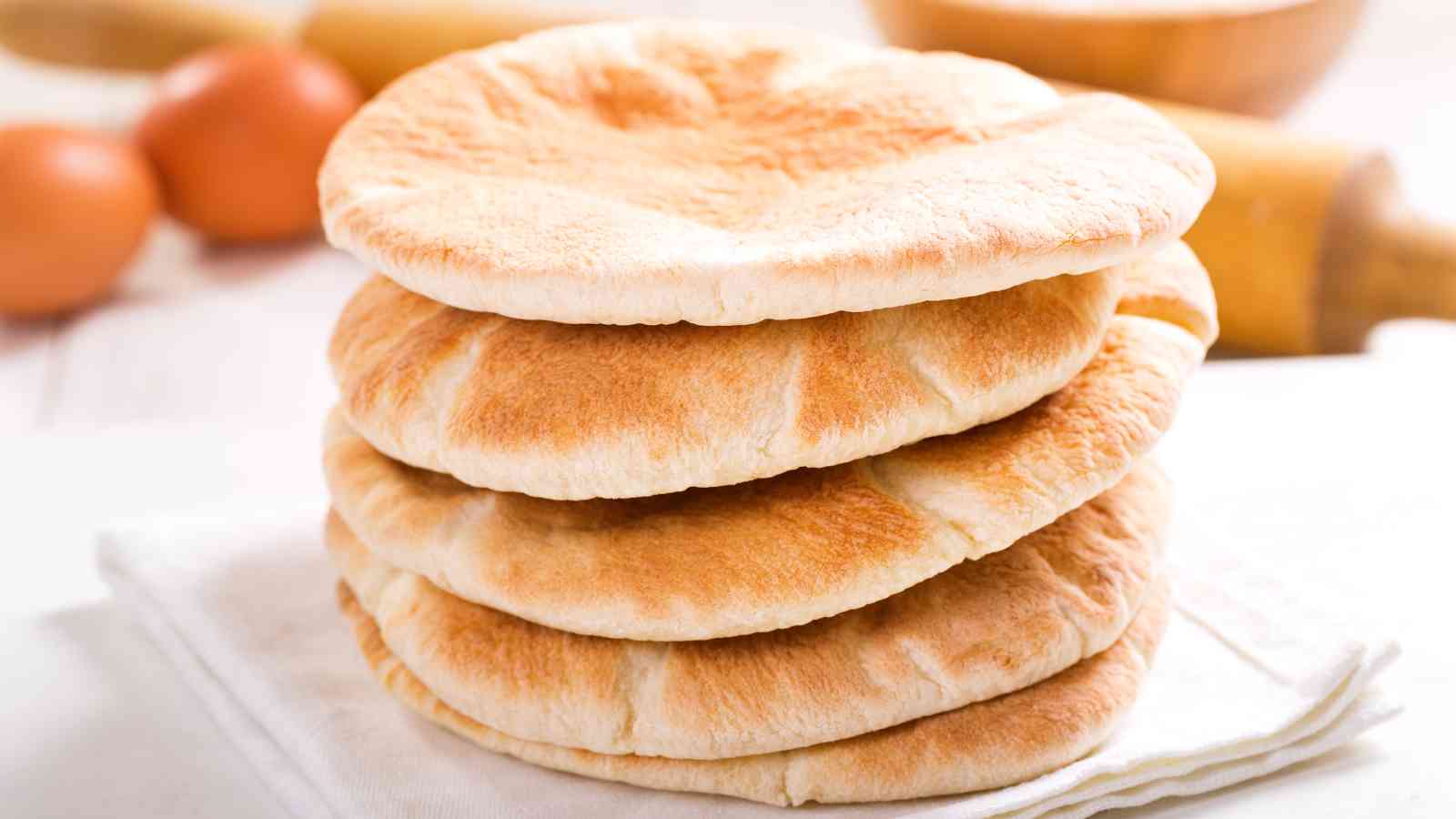 A stack of five round, golden-brown pita breads sits on a folded white cloth, with blurred kitchen items in the background.