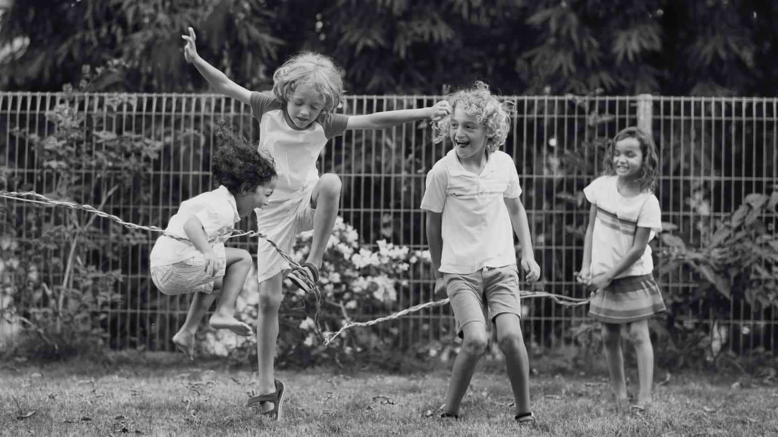 Four children play outside, with two jumping over a rope held by the others, in front of a wire fence and greenery.