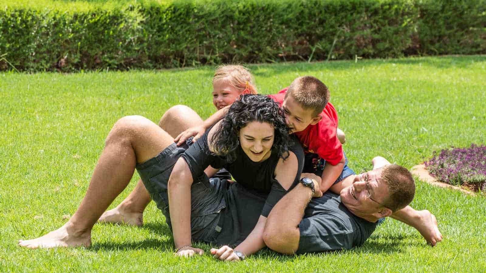A family of four, two adults and two children, playfully pile on top of each other on a grassy lawn outdoors.