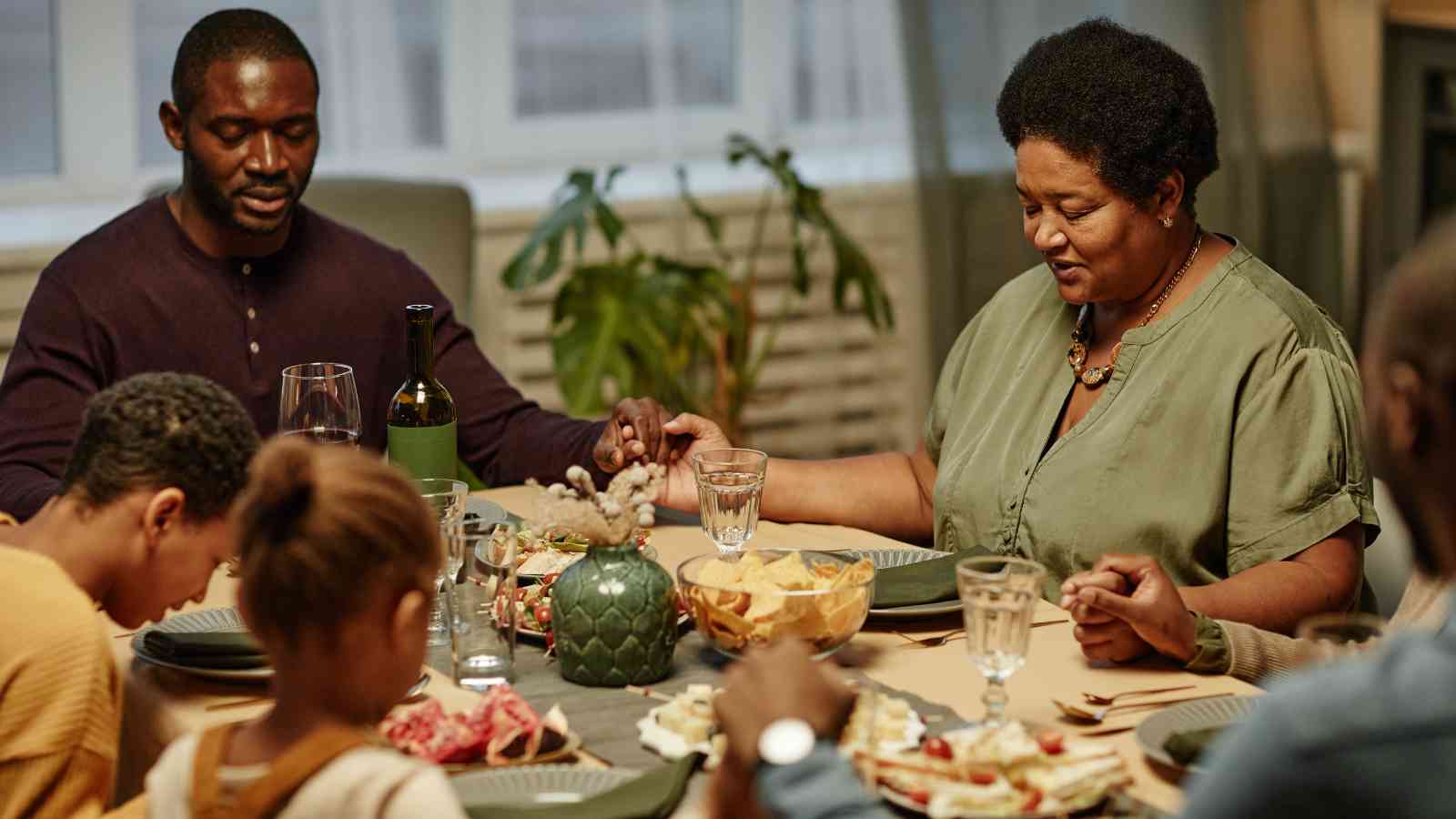 A family sits around a dining table, holding hands and praying before a meal. Various dishes and drinks are set on the table.