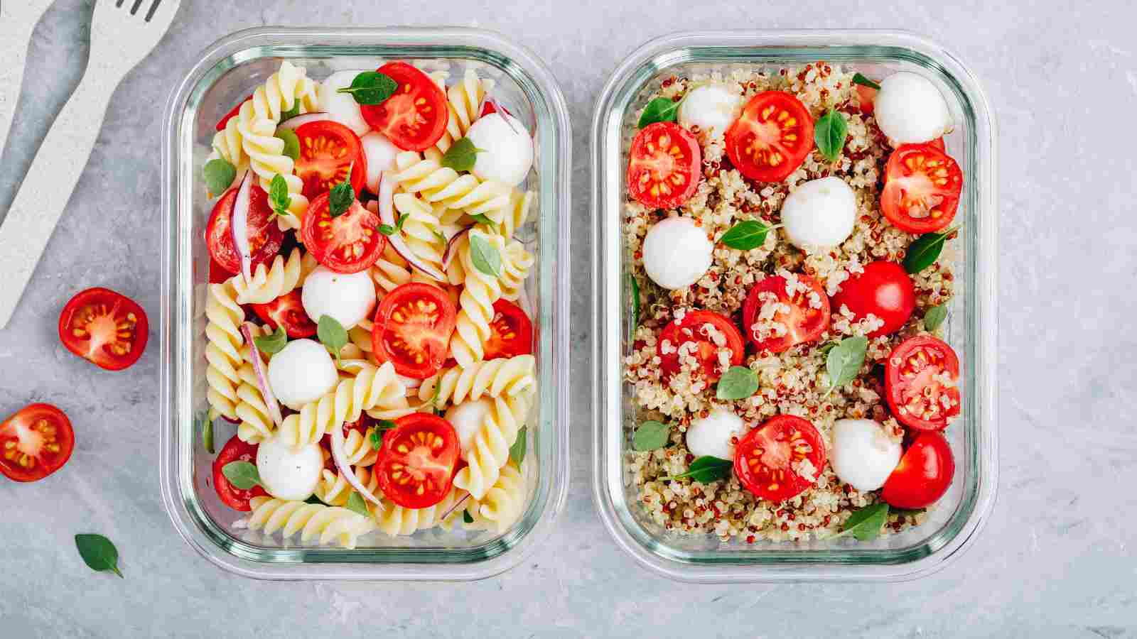 Two glass containers, one with fusilli pasta, cherry tomatoes, mozzarella balls, and basil; the other with quinoa, cherry tomatoes, mozzarella balls, and basil on a light surface.