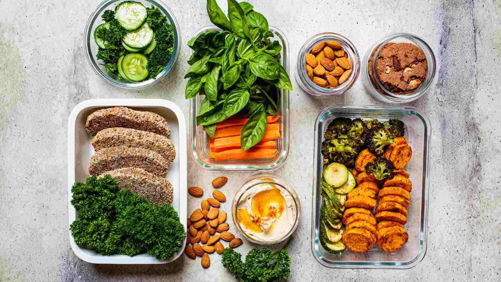 Assorted healthy foods in glass containers, including sliced bread, kale, cucumber, basil, carrot sticks, hummus, almonds, cookies, zucchini, and sweet potato.
