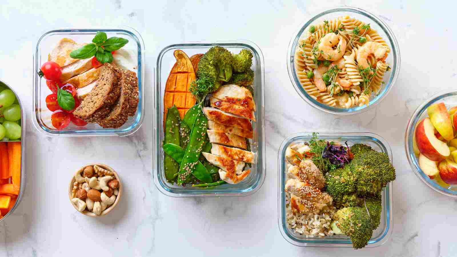An assortment of glass meal prep containers filled with various foods, including grilled chicken, vegetables, pasta with shrimp, nuts, and sliced fruit, arranged on a white surface.