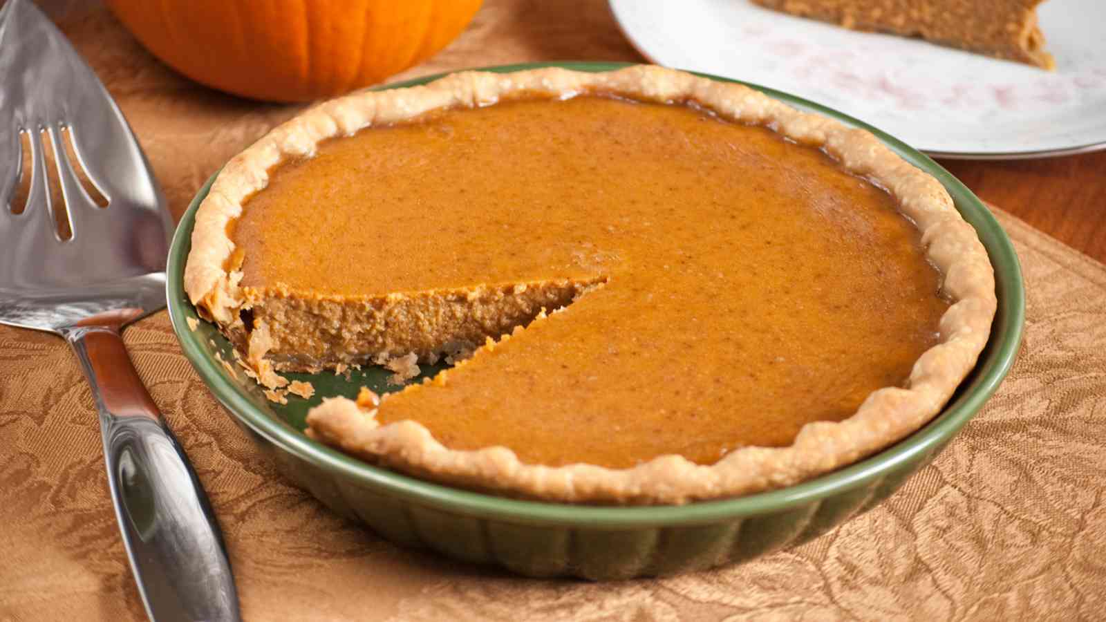 A pumpkin pie with a single slice missing, displayed in a green pie dish on a table with a serving utensil nearby.