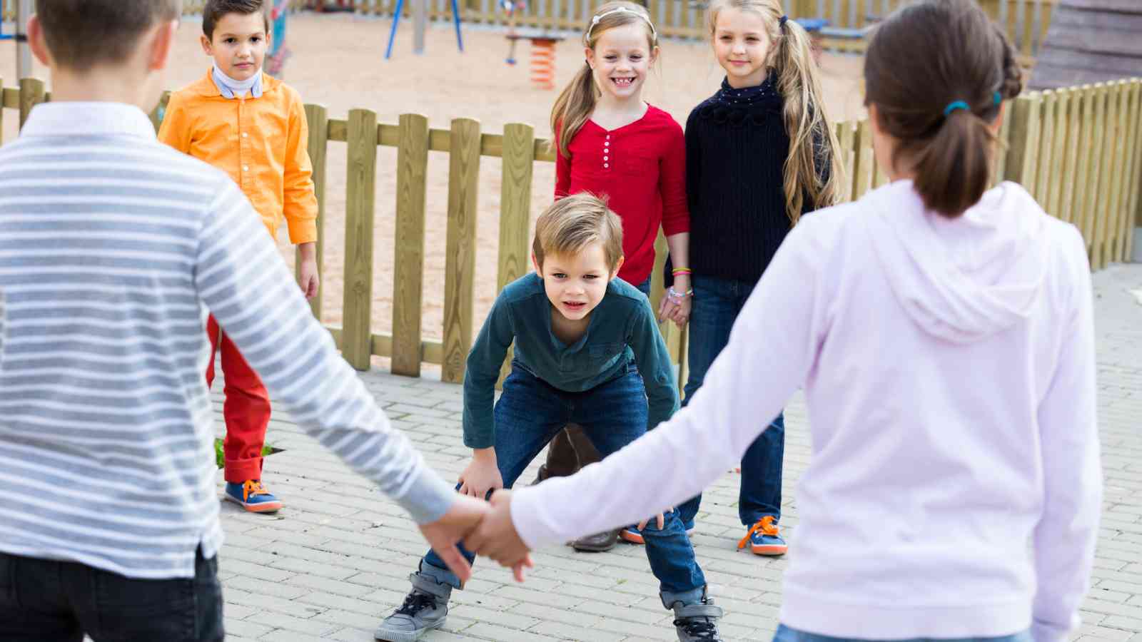 A group of children stand in a circle holding hands outdoors, while one child crouches in the center on a paved area near a playground.