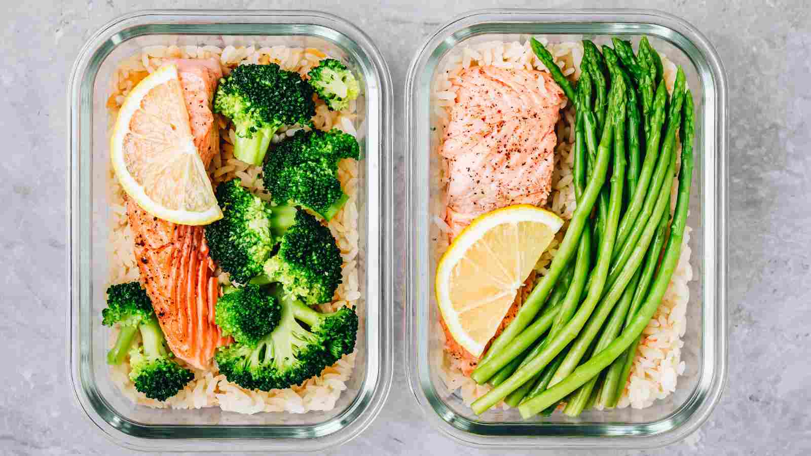 Two glass containers with rice, baked salmon topped with lemon slices, and sides of broccoli in one container and asparagus in the other, on a gray surface.