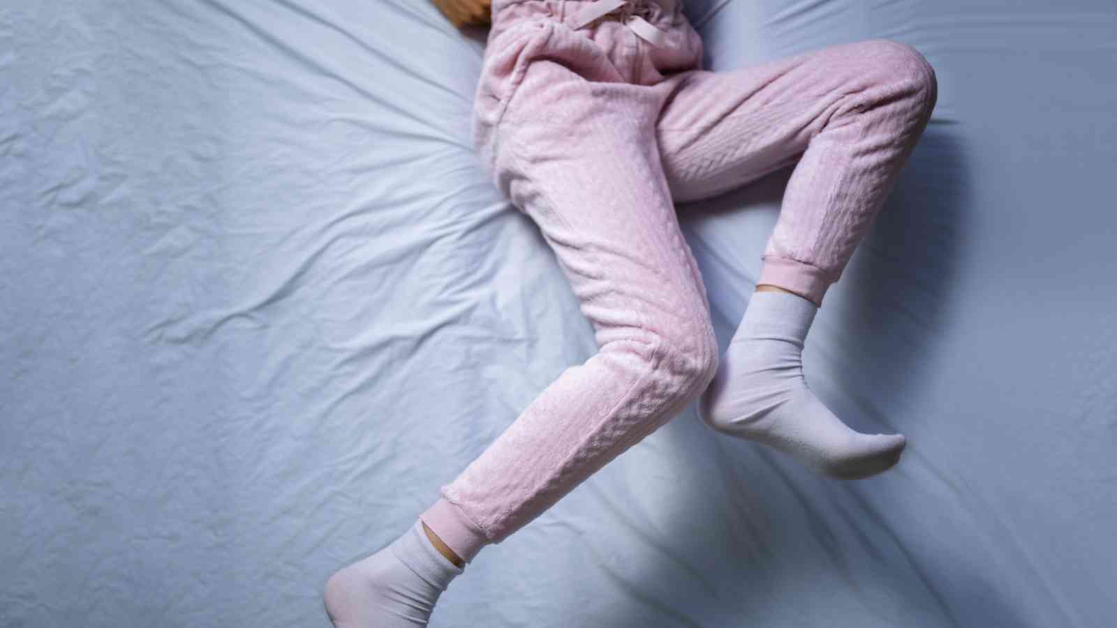 A person in light pink pajamas and white socks is lying on a bed with light blue sheets, legs slightly apart, and only the lower body visible.