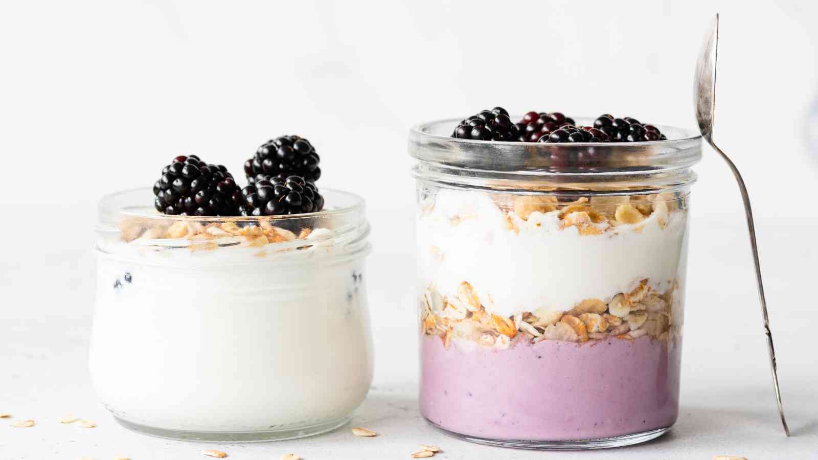 Two glass jars filled with yogurt parfaits, layered with granola and topped with blackberries, sit on a white surface. A spoon rests upright in one of the jars.