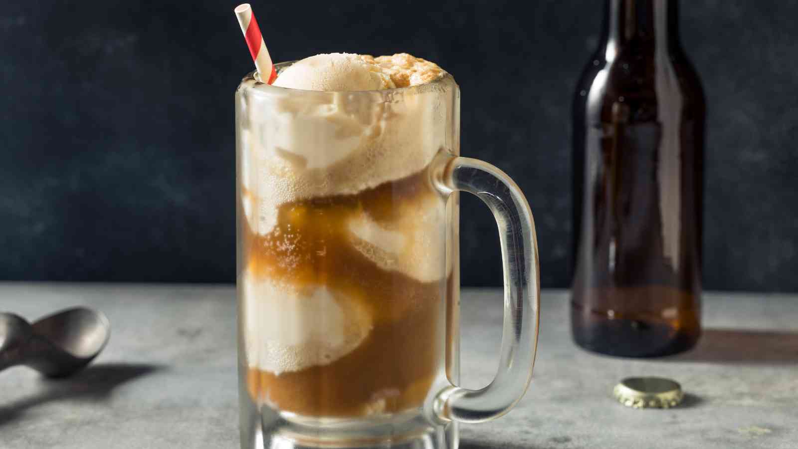 A glass mug filled with root beer and scoops of vanilla ice cream, with a red and white striped straw, sits on a gray surface beside a bottle and a bottle cap.
