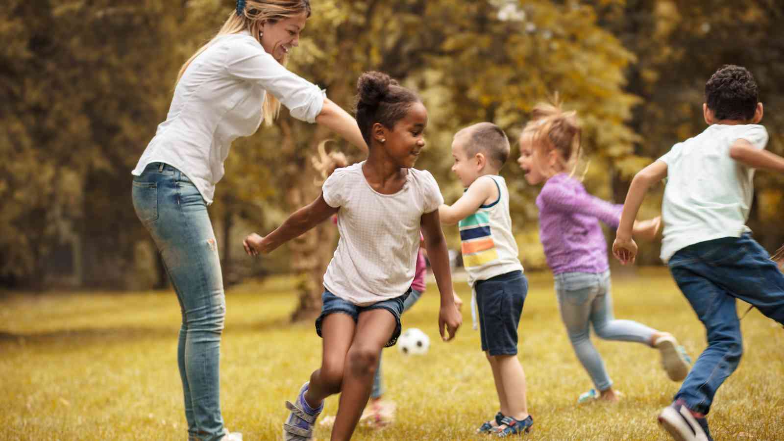 An adult and several young children play together on grass in a park, with trees in the background and a soccer ball nearby.