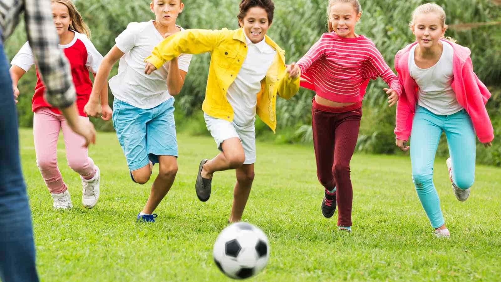 Five children running on grass chasing a soccer ball, with an adult partially visible in the foreground.