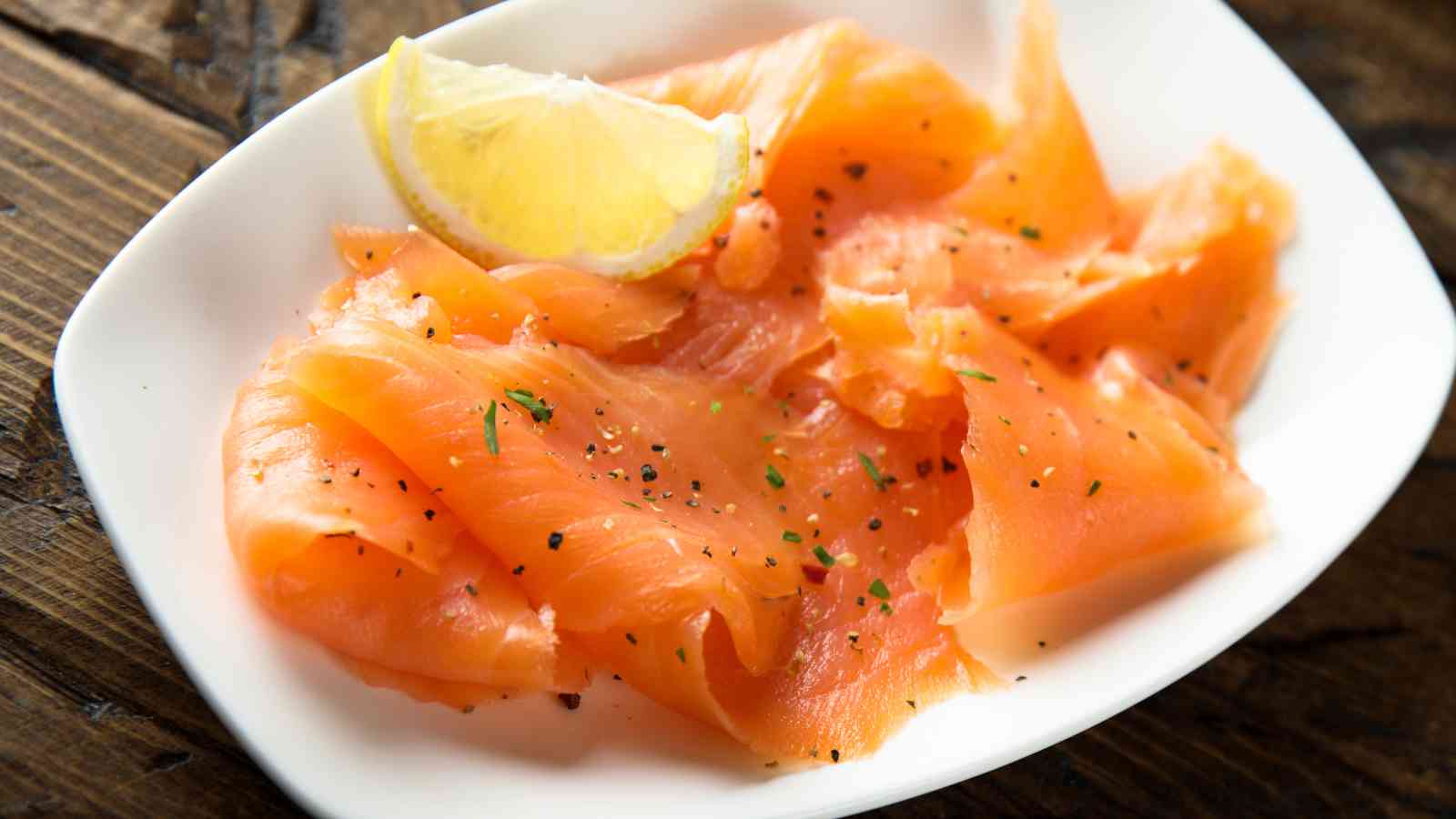 Thin slices of smoked salmon are arranged on a white plate, garnished with black pepper, herbs, and a lemon wedge.