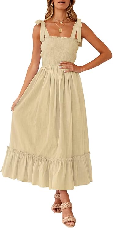 A woman wearing a light beige, sleeveless, midi dress with tie straps, smocked bodice, ruffle hem, and braided sandals.