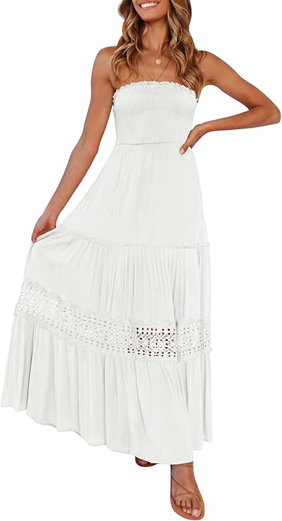 A woman wearing a strapless white maxi dress with a smocked bodice, crochet detailing, and tiered skirt, standing and holding one side of the dress.