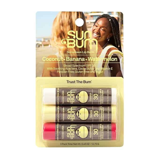 Three Sun Bum SPF 30 sunscreen lip balm sticks in coconut, banana, and watermelon flavors are displayed in yellow packaging with a smiling woman in the background.