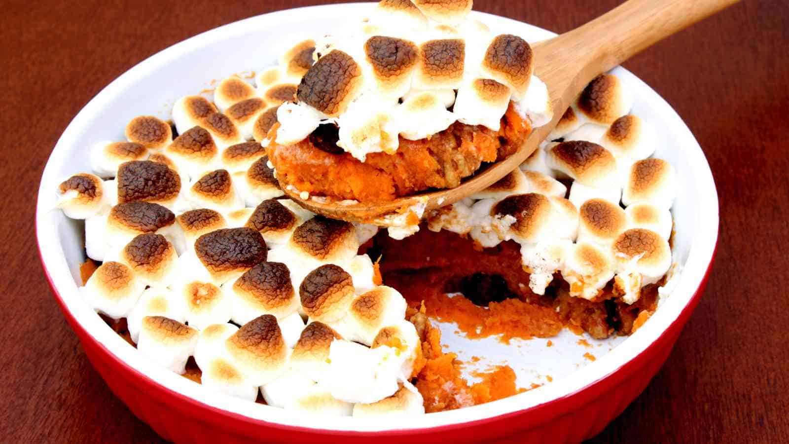 A casserole dish of sweet potato topped with toasted marshmallows, with a portion being served using a wooden spoon.