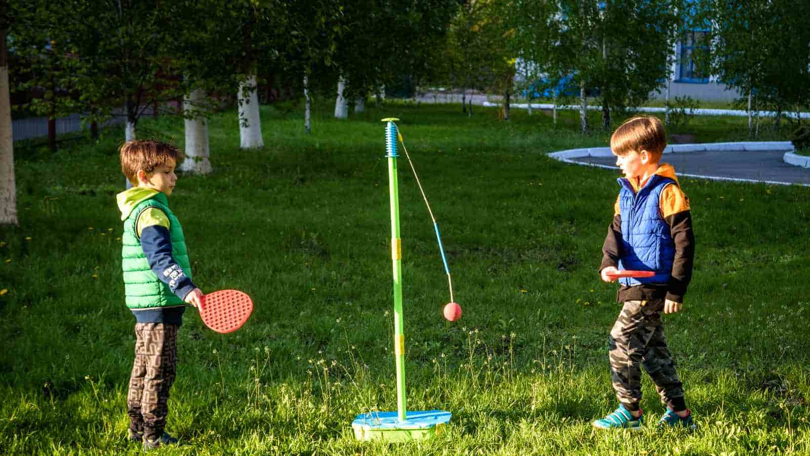 Two boys stand on grass and play tetherball with red paddles and a ball attached to a pole in a park setting with trees in the background.