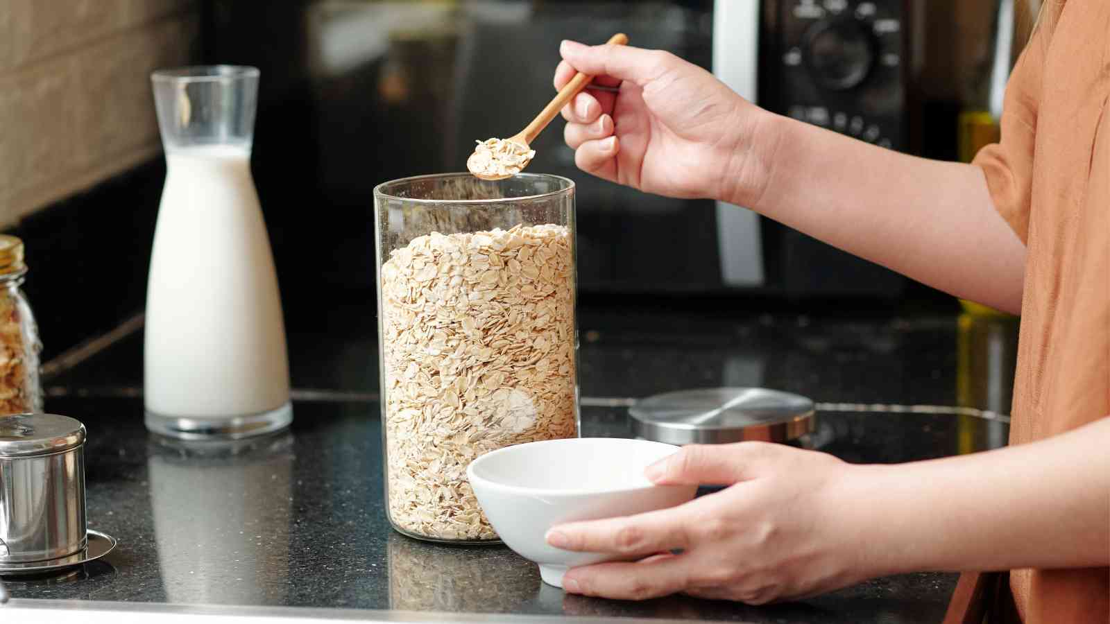 A person scoops rolled oats from a glass container into a white bowl, with a glass pitcher of milk on the counter nearby.