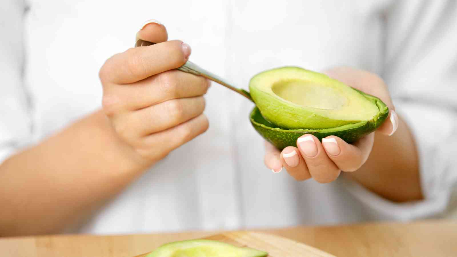 A person scoops avocado flesh from a halved avocado with a spoon, holding the fruit in one hand over a wooden surface.