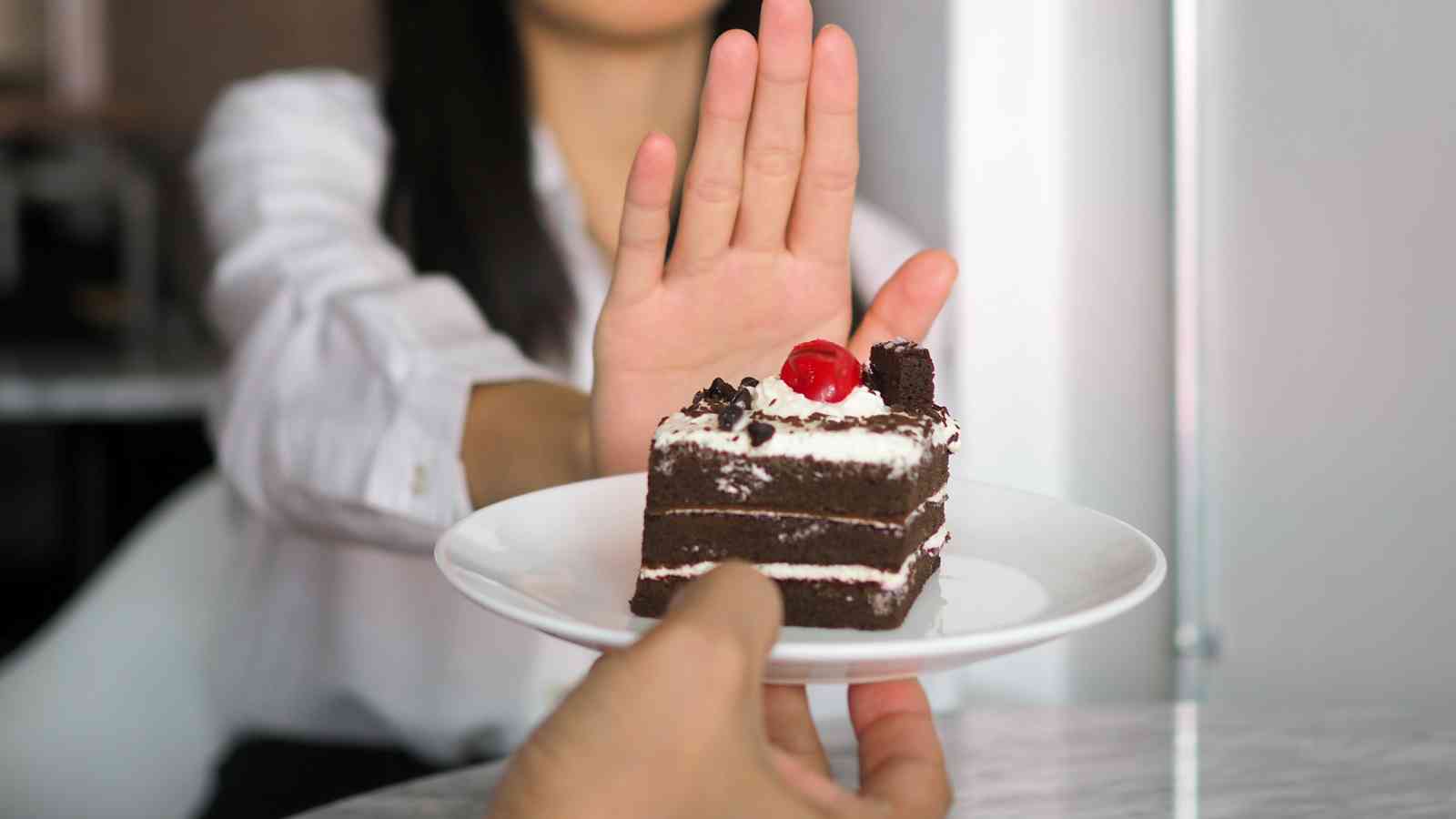 A person holding up their hand to refuse a plate of chocolate cake with a cherry on top offered by another person.