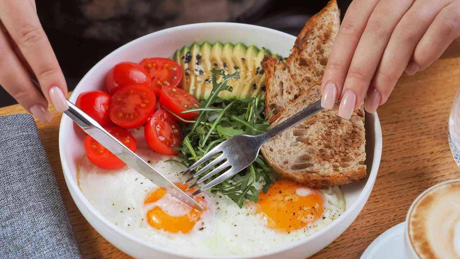 A person cuts into fried eggs in a bowl with cherry tomatoes, avocado slices, arugula, and bread on a wooden table.