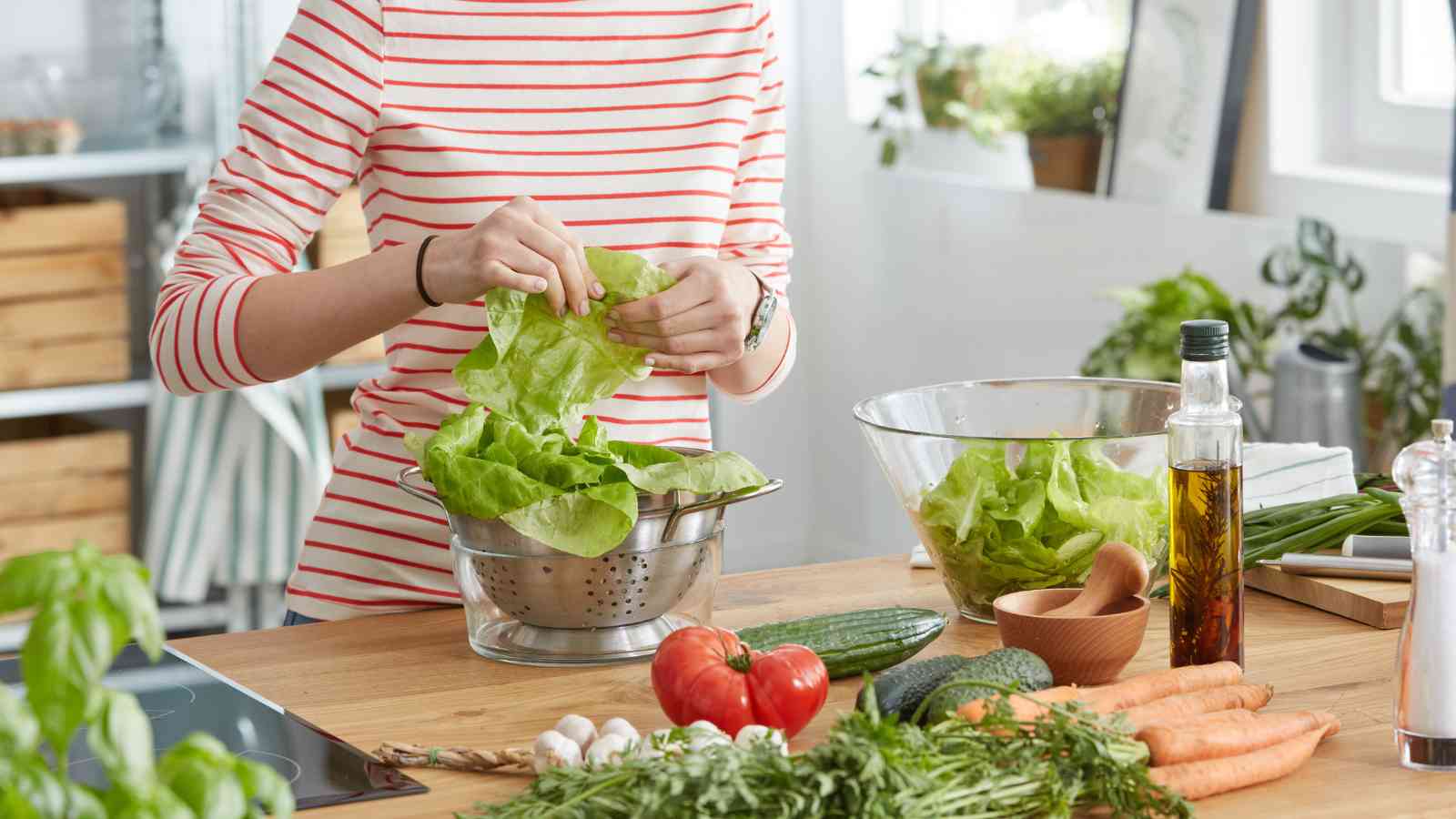 Person preparing a salad in a kitchen, tearing lettuce into a colander surrounded by fresh vegetables, olive oil, and kitchenware on a wooden countertop.