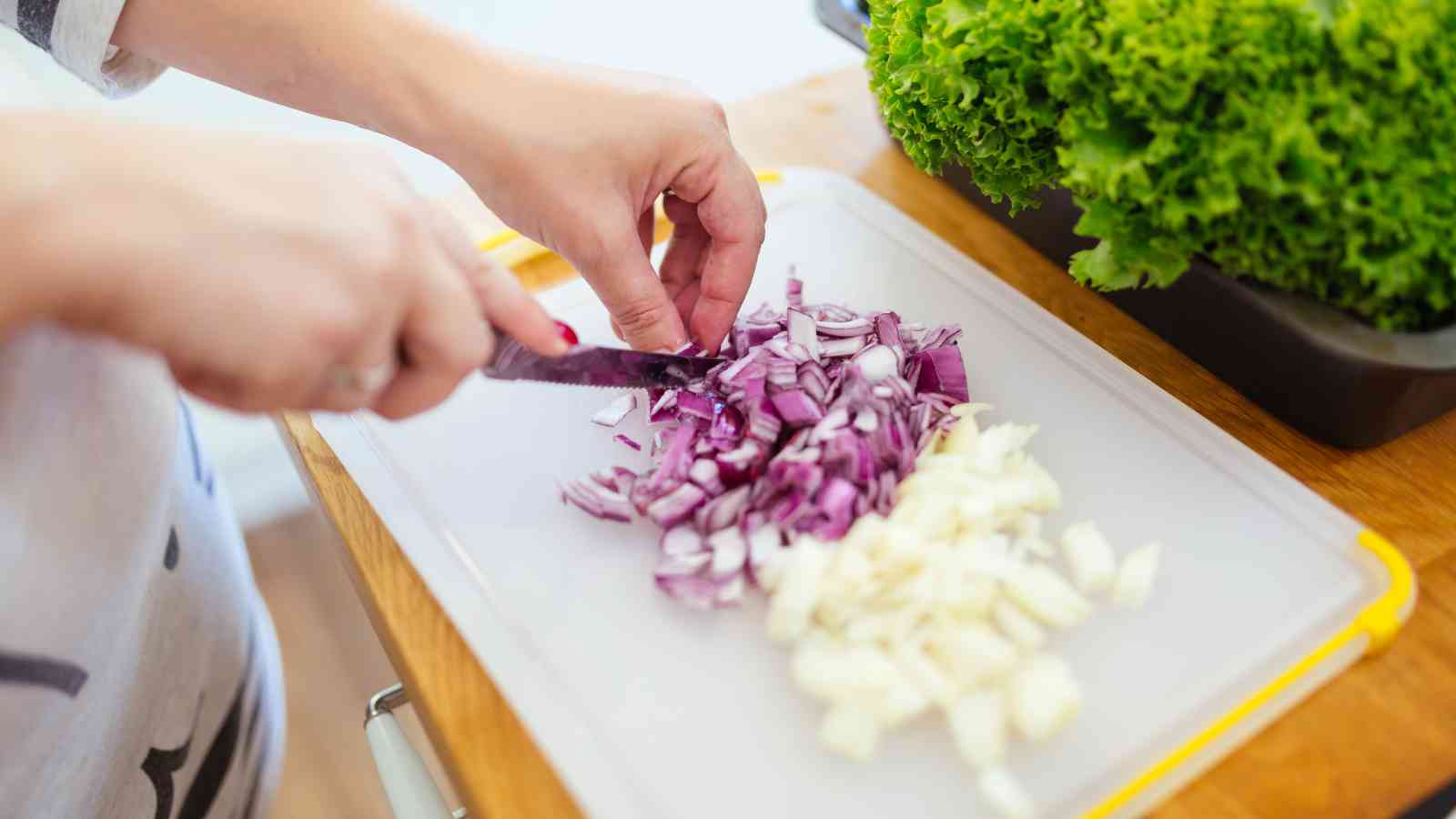 A person chopping red onions and garlic on a white cutting board with fresh green lettuce nearby.