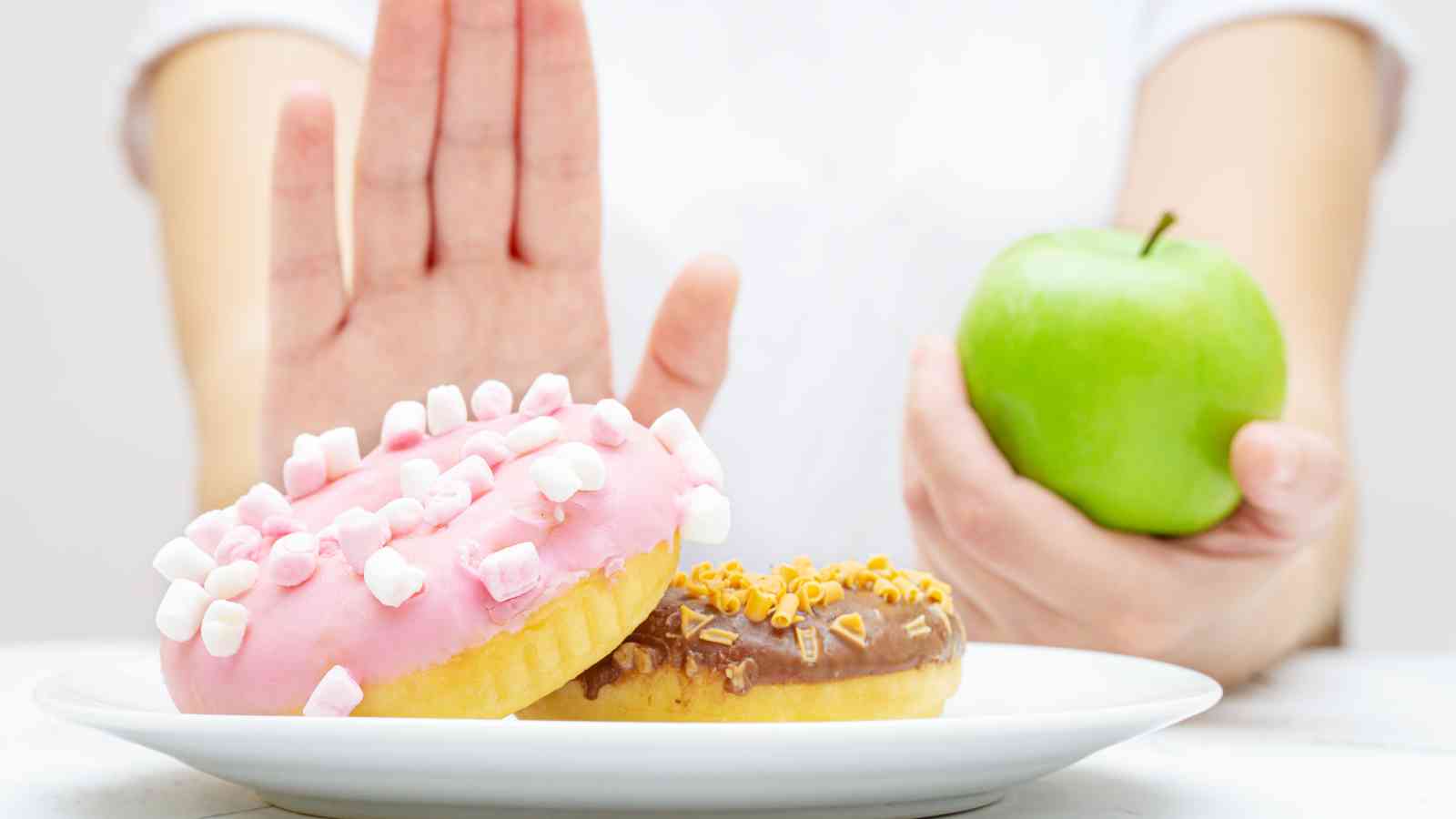 A person holds a green apple in one hand and raises the other hand to refuse a plate of doughnuts topped with pink and chocolate icing.